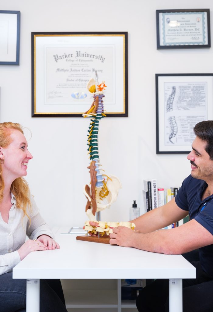 Chiropractor en patiënt in gesprek aan bureau met wervelkolommodel in de spreekkamer van de praktijk
